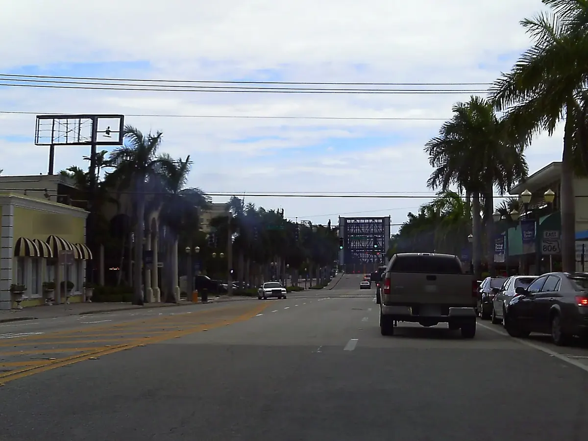 Palm-lined Atlantic Avenue in downtown Delray Beach, with a pickup truck and parked cars on the way to the drawbridge